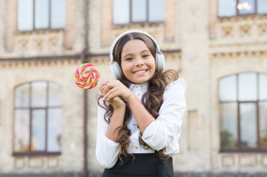 Tatlı onu mutlu ediyor. Okul kızının modern görünüşü. Kulaklıklı çocuk lolipop tutuyor. Öğrenci müzik dinler. Okula dönüyorum. Şekerli güzellik. Çocuk eğitimde yenilik yapıyor. Mutlu çocuklar günü. Benim tatlı hayatım