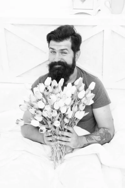 Make surprise concept. Gift for spouse. Bearded hipster in bed. Spring in bedroom. Man hold tulips bouquet while relaxing in bed. Fresh flowers. Flowers delivery service. Birthday anniversary holiday