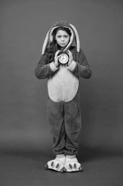 Morgonlarm. Regelbunden dag börjar. Förtjusande kanin hålla väckarklocka. En liten flicka i kanindräkt. Barnkaninkigurumi. En kaninpyjamas. Bunny kid röd bakgrund. Bebisdjur. Räknetid — Stockfoto