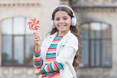Küçük kız şeker yiyor. Havalı hippi kıyafetleri içinde mutlu bir kız öğrenci. çocukluk mutluluğu. Dijital kulaklıklı çocuk. Küçük kız lolipopla müzik dinliyor. Okula geri dön.