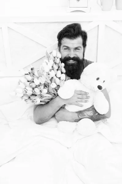 Teddy bear. Fresh flowers. Bearded hipster in bed. Valentines day gift. Birthday holiday. Man hold tulips bouquet relaxing in bed. Flowers delivery service. Make surprise concept. Gift for spouse
