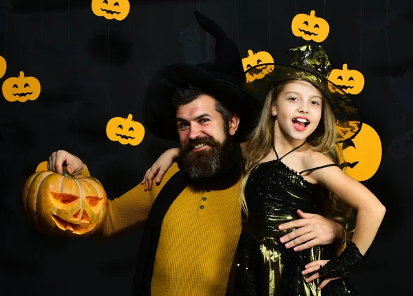 Wizard and little witch in black hats hold pumpkin.