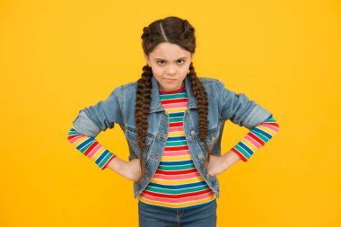 Kötü bir mizah içinde. Huysuz küçük çocuk sarı arka plan. Küçük bebek örgülü saç stili giyer. Sıradan tarzda küçük bir kız. Elleri kalçasında küçük bir çocuk. Moda. Şık trendler. Kuaför salonu.