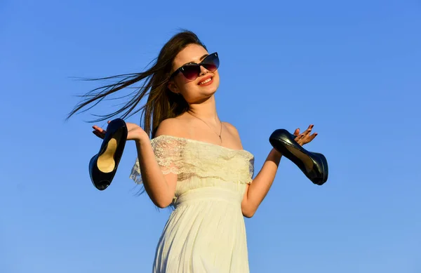 Mujer en el soleado cielo azul de la mañana. Vacaciones de verano. Modelo tierno vestido de verano. Libertad. Chica en gafas de sol copiar espacio. Hacia el verano. Concepto post fiesta. Mujer amanecer llevar tacones altos en la mano — Foto de Stock