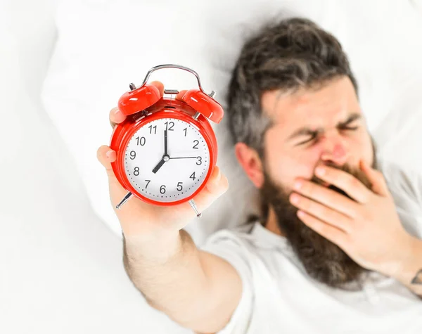 Man with yawning face lies on pillow, holds alarm clock.