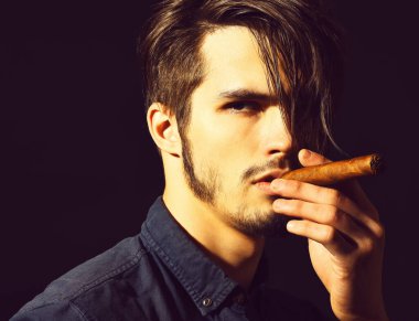 Ciddi sakallı bir adam siyah stüdyo arka planında puro içiyor.