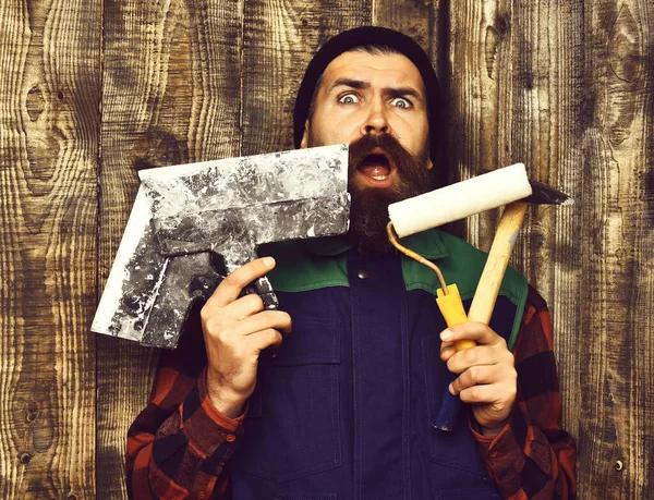 Homem construtor barbudo segurando várias ferramentas de construção com rosto surpreso — Fotografia de Stock