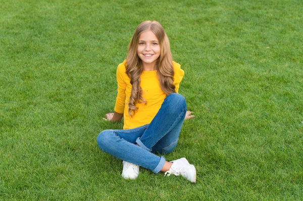 Menina feliz tem cabelo encaracolado longo. criança beleza e moda. menina adolescente após cabeleireiro. relaxar na grama verde. tempo de lazer primavera. infância feliz. criança com cabelo elegante. sorriso alegre — Fotografia de Stock