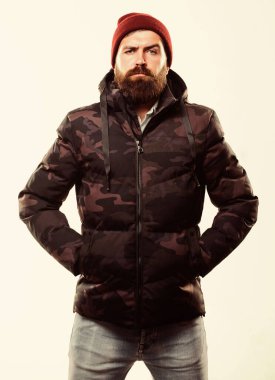 Rahat bir kış kıyafeti. Adam beyaz arka planda kapüşonlu kalın kamuflaj desenli ceket giyiyor. Şapka takan ve siyah kışlık ceket giyen adam. Hipster kış modası. Kış stili erkek giyim