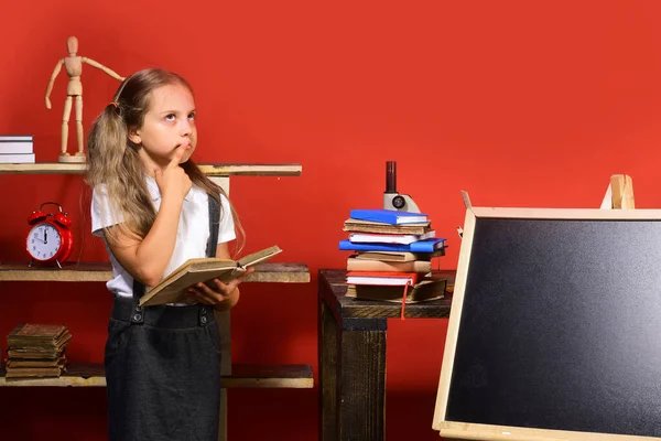 Girl holds open book and stands by blank blackboard — Stock Photo, Image