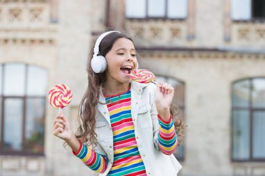 Gelecekteki şarkıcı. Dijital kulaklıklı çocuk. Küçük kız lolipopta müzik dinliyor. Okula dönüyorum. Küçük kız şeker yiyor. Havalı hippi kıyafetleri içinde mutlu bir kız öğrenci. çocukluk mutluluğu