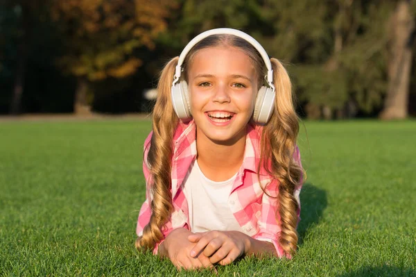 音楽を聴いてる。学校に戻って。公園で勉強してる。ヘッドフォンで緑の草の上でリラックス。小さい女の子はオーディオブックを聞く。子供のための新技術。幸せな子供の頃の記憶。今日学ぶ. — ストック写真