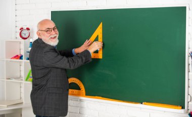Gözlüklü hoca tahtada üçgen çiziyor. Okula dönüyorum. Hangi açıdan bakıyorsun? Geometrik şekiller. Lise çağdaş eğitim. Usta öğretmen çizim yaparken üçgen kullanıyor.