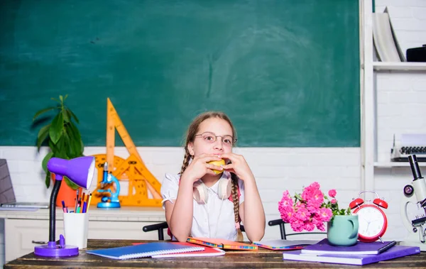 Svačinka mezi lekcemi. Školačka sedět stůl tabuli pozadí. Student na škole. Holka, co jí jablečný ovoce. Školní životní koncept. Zdravý životní styl. Moderní dětství. Školní přestávka — Stock fotografie