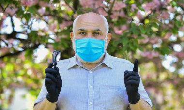 Kurallara uy. Tıbbi maske ve eldiven giymiş kıdemli bir adam. Parktaki sakura ağacı. Coronavirus kapanışı sırasında hayat. Hastalık korkusu. Virüsle savaşma stratejisi. Halka açık yerlerde ziyaret yasağı