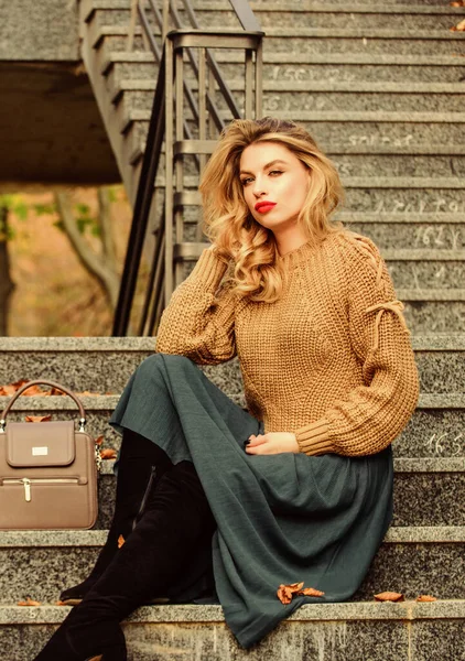 How to Style Sweater and Skirt Combo for Fall. Woman with gorgeous hairstyle sit on stairs outdoors. Fall fashion trend. Layer oversize knit over girly skirt. Wearable trends. Fall outfit formula