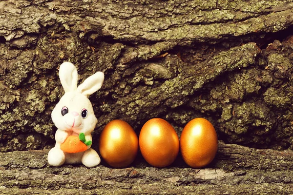 Jouet de lapin, oeufs de Pâques dorés sur fond d'écorce d'arbre — Photo