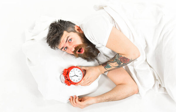 Man with shocked face lies on pillow near alarm clock.