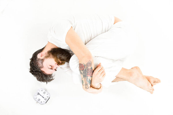 Man in white pajama with sleepy face lies near clock.