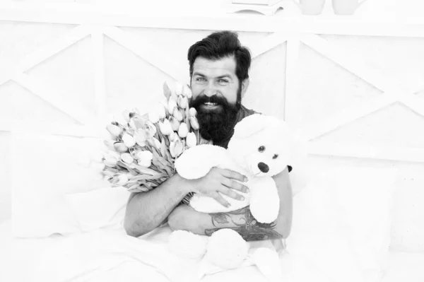 Cute teddy bear. Fresh flowers. Bearded hipster in bed. Valentines day gift. Birthday holiday. Man hold bouquet relaxing in bed. Flowers delivery service. Make surprise concept. Gift for spouse