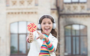 Üzerinde lolipop olan kablosuz kulaklıklı küçük bir kız. Okula dönüyorum. Yeni teknolojiyle modern eğitim. Çocuk okul bahçesinde şarkı söylüyor. Sağlıklı yemek. Çocuğun müzik molası var. Kulaklıklı kız.