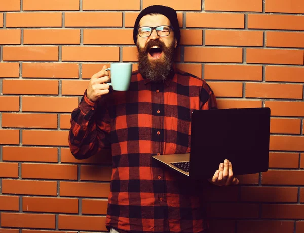 Bearded brutal caucasian hipster holding laptop with cup — Stock Photo, Image