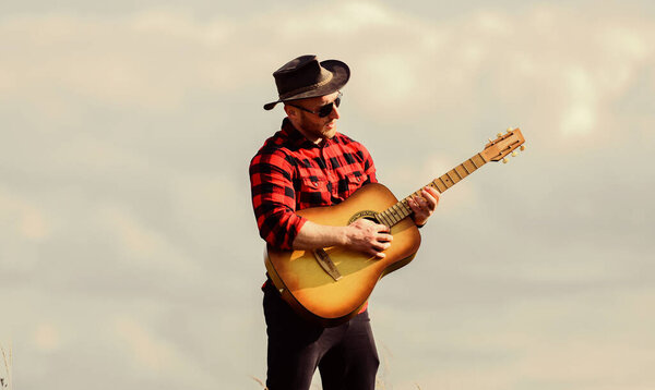 Hiking song. Handsome man with guitar. Country style. Summer vacation. Play beautiful melody. Country music concept. Guitarist country singer stand in field sky background. Inspired country musician