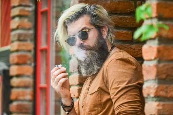 Smoking outdoors. Went on smoke break. Hipster smoking old architecture background. Smoking habit. Fashionable mature man with cigarette. Brutal guy sunglasses smoking tobacco. Cool guy relaxing