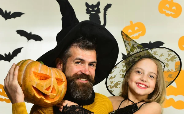 Wizard and little witch in black hats hold pumpkin.