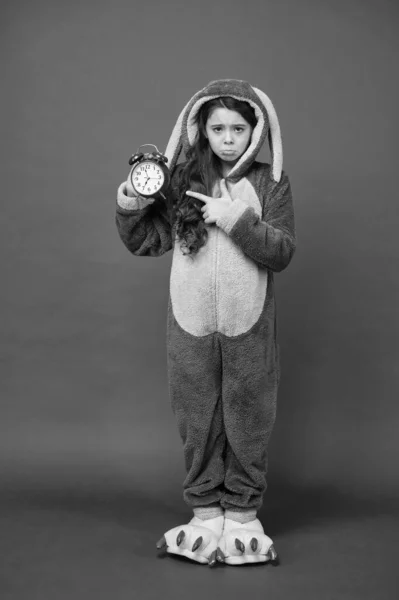 Δυστυχισμένο παιδί στο κιγκουρούμι πιτζάμα να κρατάει το ξυπνητήρι. Ώρα να ξυπνήσεις. Όνειρα γλυκά Καλημέρα και σε σένα. Άνθρωποι και ιδέες για ύπνο. Το κοριτσάκι φοράει στολή πασχαλινού λαγού. Νωρίς σηκώθηκες. διαχείριση του χρόνου — Φωτογραφία Αρχείου