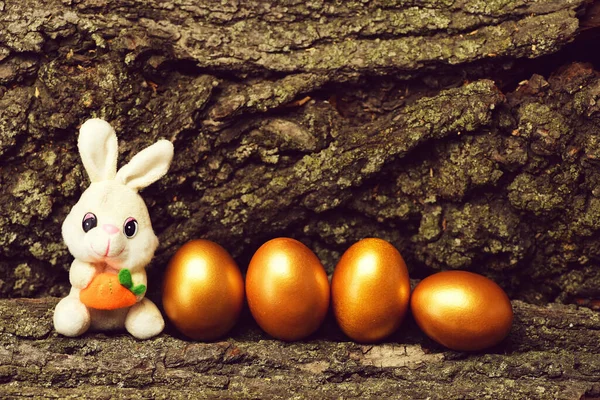 Jouet de lapin, oeufs de Pâques dorés sur fond d'écorce d'arbre — Photo