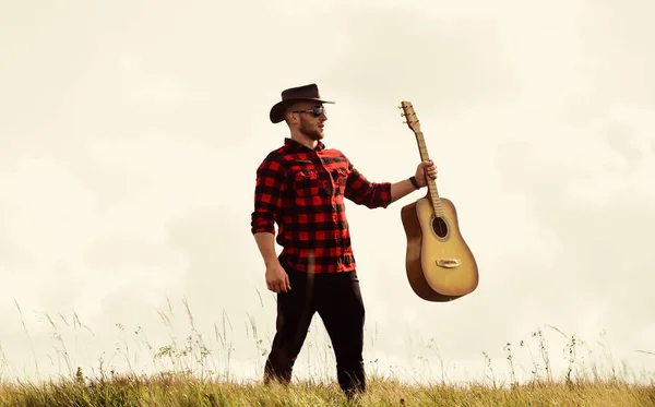 Concepto de música country. Guitarrista cantante country de pie en el fondo del cielo de campo. Estilo country. Vacaciones de verano. Canción de senderismo. Músico country inspirado. Unidos con la naturaleza. Hombre guapo con guitarra — Foto de Stock