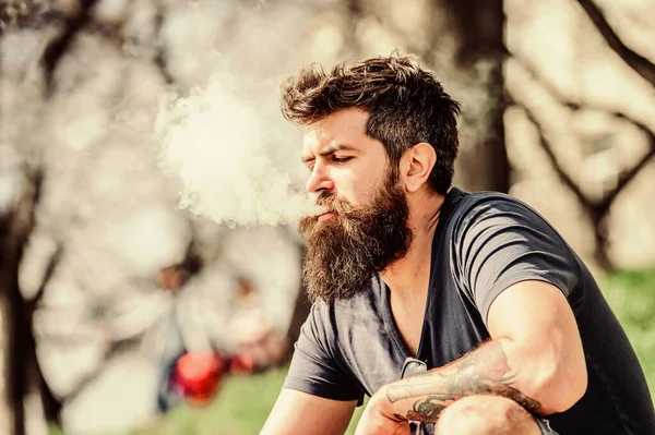 Man long beard relaxed with smoking habit. Man with beard and mustache breathe out smoke. White clouds of flavored smoke. Stress relief concept. Bearded man smoking vape. Smoking electronic cigarette