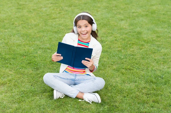 Pracuj tvrdě. Malá holčička čte knihu. literatura pro děti. psát vzpomínky z dětství. chytrý kluk ve sluchátkách. Zpátky do školy. děti studují online v parku. poslouchat hudbu na zelené trávě s knihou — Stock fotografie