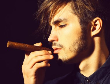 Ciddi sakallı bir adam siyah stüdyo arka planında puro içiyor.