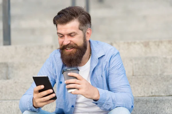 Comunicación en línea de la vida moderna. Tipo con fondo de espacio urbano smartphone. Hombre barbudo guapo relajante teléfono móvil y taza de café. Tecnología moderna. Vivir la vida en línea. Redes sociales — Foto de Stock