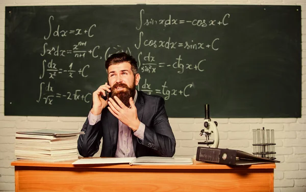 Calling parents. School teacher call mobile phone while sit classroom chalkboard background. Teacher bearded man talk mobile phone. Call colleague ask advice. Pedagogue keep in touch with colleagues