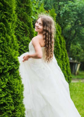Yakışmış. Kadın kuaför tarzı. Uzun kıvırcık saçlı küçük bir kız. Beyaz elbiseli küçük bir güzellik. düğün meleği çocuk. Çocuk, thuja ile park etmekten hoşlanıyor. bahar selvi ağacı. Yaz doğası