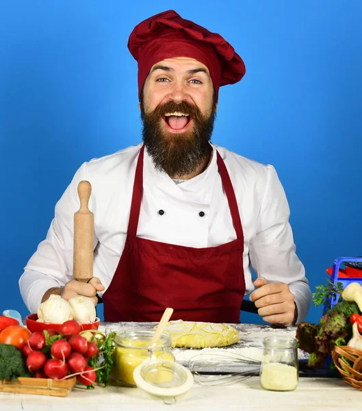 Cook with cheerful face in burgundy uniform holds rolling pin. — Stock Photo, Image