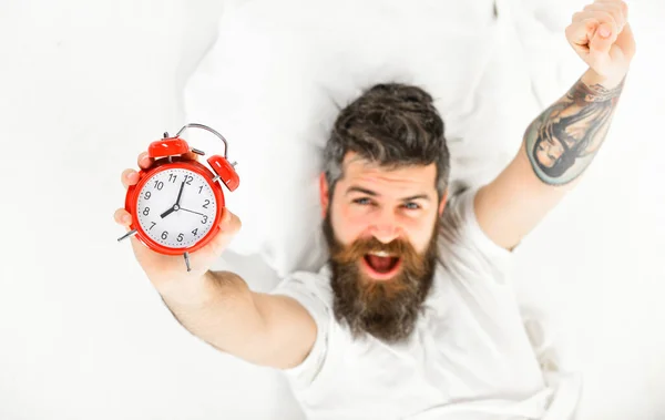 El hombre con cara de bostezo feliz yace en la almohada, sostiene el reloj . —  Fotos de Stock