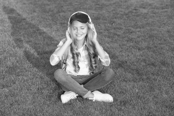 屋外でリラックスしながら音楽を聴く。子供の女の子は音楽の緑の草の牧草地を楽しむ。楽しい時間だ。子供のヘッドフォンは音楽を聞く。女の子のヘッドフォン音楽を聞いて。教育用ポッドキャスト。いつでもお楽しみください — ストック写真