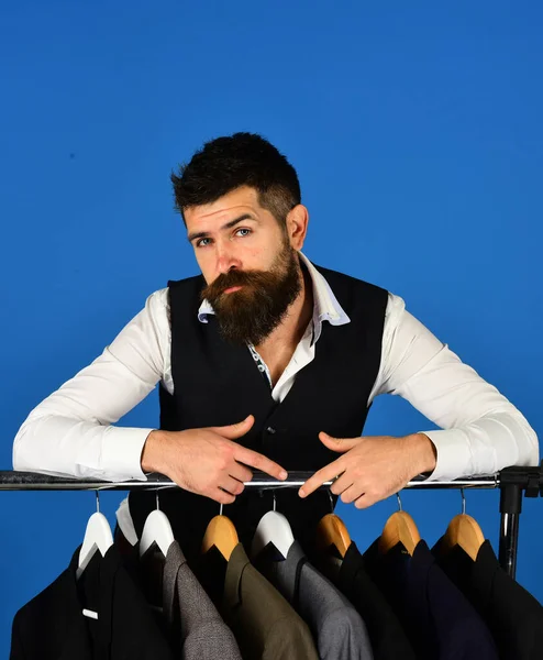 Man with beard by clothes rack. Designer leans on clothes