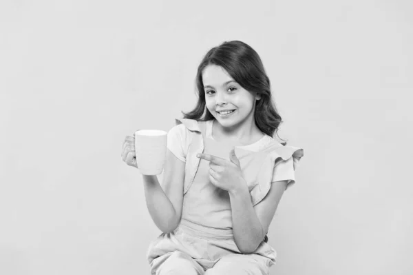 No use crying over spilt milk. Drink enough water. Girl kid hold mug yellow background. Carefree smiling child pointing at mug. Drinking tea juice cocoa. Relaxing with drink. Child drink beverage.