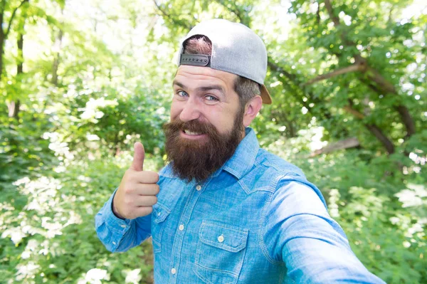 Bienvenidos a nuestra fogata. chico maduro en sombrero. feliz viajero hipster. hacer selfie en el bosque al aire libre con la naturaleza puesta del sol en el fondo. Barbudo estilo casual hombre. retrato de moda del hombre barbudo — Foto de Stock