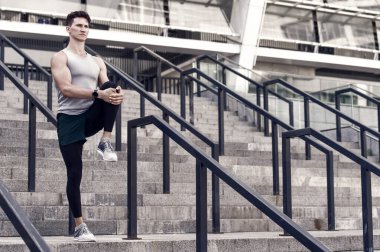 Spor giysileri içinde koşan bir sporcunun portresi. Genç fitness koşucusu koşmadan önce bacaklarını esnetiyor. İlk önce ısınalım. Sağlıklı yaşam ve kendini kontrol kavramı. Adam esniyor.