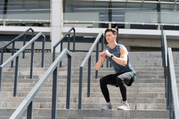 Sporten en een gezonde levensstijl concept. Man doet kraakpanden buiten. Een man met een fitnesshorloge die traint en squats doet. Diepe kraakpijnen. jongeman in sportkleding doet hurken buiten — Stockfoto