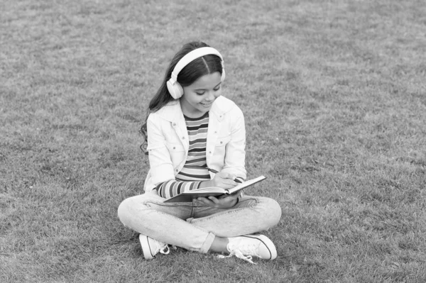 写歌歌词。快乐童年的概念。孩子把记忆写在日记里。女学生拿着笔记本放松。快乐的孩子在耳机里听音乐。在绿草上的小女孩。孩子们喜欢公园自然的歌声 — 图库照片
