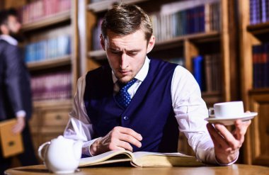 Genç adam elinde çaydanlık ve kitapla masanın arkasında oturuyor.