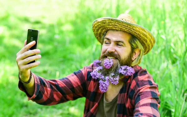Homem maduro jardineiro relaxar na grama verde. desfrutar da natureza primavera. selfie verão com lilás no telefone. rancho brutal hipster tem longa barba saudável. flor no cabelo. agricultor relaxado em chapéu de palha — Fotografia de Stock