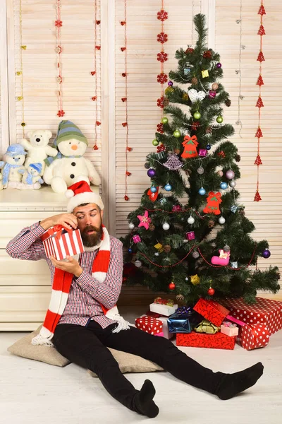 Guy in hat and scarf sits by Christmas tree. — Stock Photo, Image